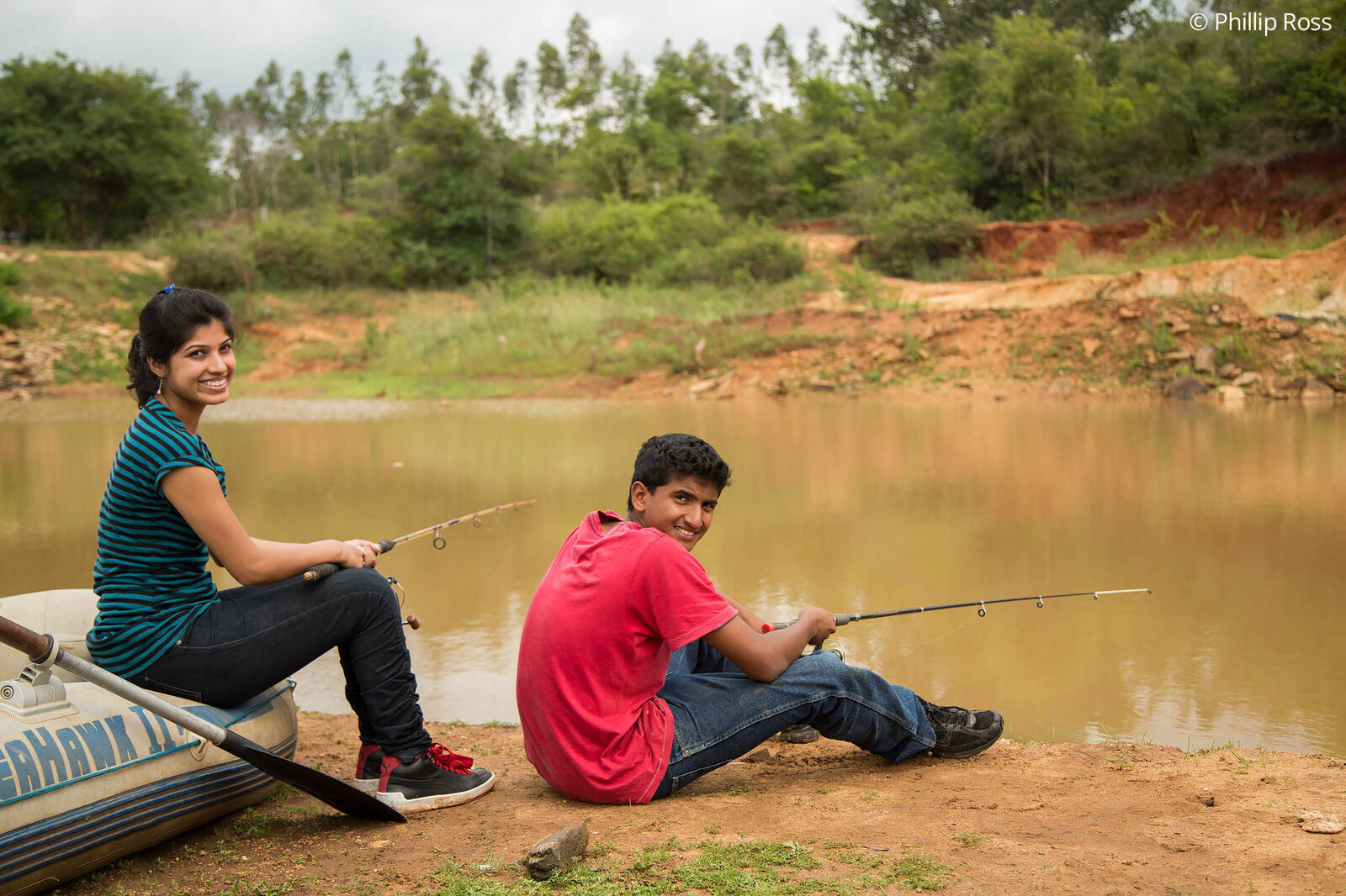 Two young guests sitting at the edge of the lake with fishing rods, beside an inflatable boat