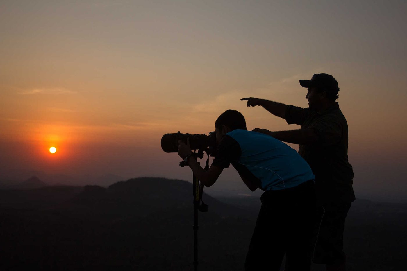 Two figures silhouetted against the setting sun, one at a long lens on a tripod and one pointing out a subject