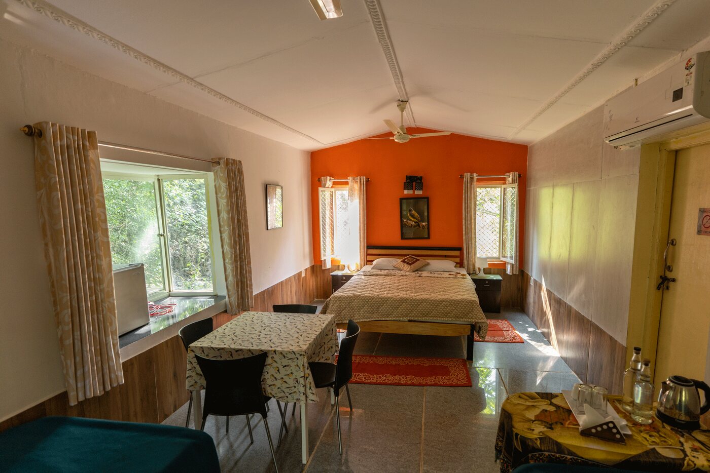 Inside the forest facing room: a queen-size bed against an orange wall, a dining table and a window onto the trees